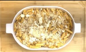 Baked ziti with cheese topping in a white dish on a wooden board, ready for serving.